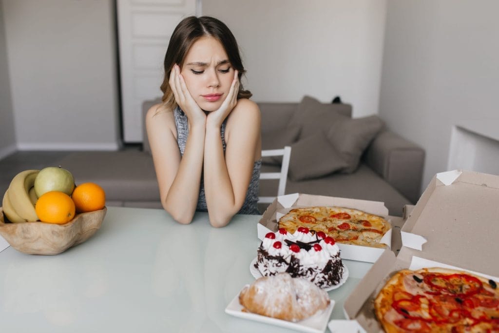 Γυναίκα με τα χέρια στο πρόσωπό της και προβληματισμένο βλέμμα κοιτάζει πίτσες και γλυκά πάνω στο τραπέζι και στην άλλη πλευρά του τραπεζιού υπάρχει ένα ξύλινο μπολ με φρούτα